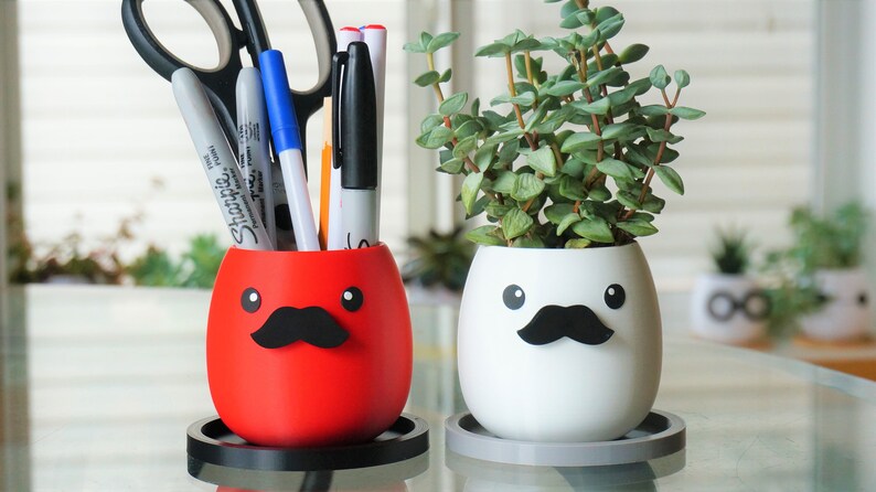 May include: Two white ceramic planters with black mustaches and googly eyes. One planter is red and holds pens and scissors. The other planter is white and holds a small green plant.