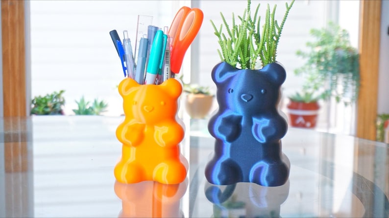 Puede incluir: Dos macetas con forma de osito de goma, una naranja y otra azul. La maceta naranja est&aacute; llena de bol&iacute;grafos y l&aacute;pices. La maceta azul tiene una peque&ntilde;a planta verde creciendo dentro.