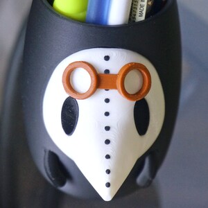 May include: Black and white ceramic pencil holder shaped like a face with orange goggles. The holder has a long nose and dotted lines around the eyes.