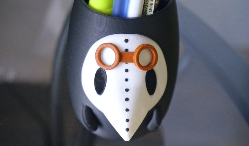 May include: Black and white ceramic pencil holder shaped like a face with orange goggles. The holder has a long nose and dotted lines around the eyes.