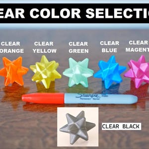 May include: A selection of clear plastic star-shaped ornaments in various colors, including red, orange, yellow, green, blue, magenta, white, and black. The text "CLEAR COLOR SELECTION" is at the top of the image. The text "CLEAR BLACK" is below the black ornament.