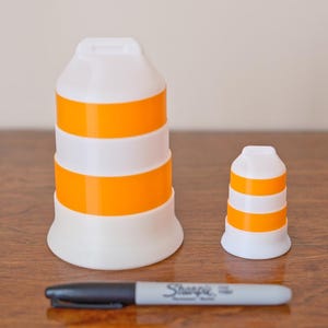 Może przedstawiać: Dwa biało-pomarańczowe, cylindryczne obiekty w paski, jeden duży i jeden mały, stoją na drewnianej powierzchni. Czarny i szary marker Sharpie leży przed nimi. Obiekty mają kształt pachołka drogowego.