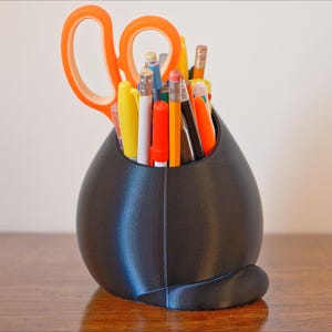 May include: A black, egg-shaped desk organizer holding various pens, pencils, and scissors. The scissors have orange handles. The organizer sits on a wooden surface, with a light-colored wall in the background.