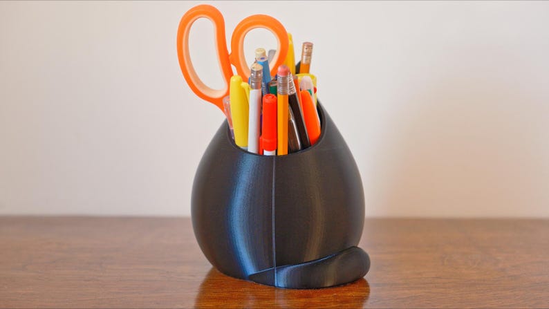 May include: A black, egg-shaped desk organizer holding various pens, pencils, and scissors. The scissors have orange handles. The organizer sits on a wooden surface, with a light-colored wall in the background.