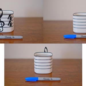 May include: A white pen holder with black horizontal lines and a treble clef design. A blue Sharpie pen rests in front of the holder. The pen holder is on a wooden surface.