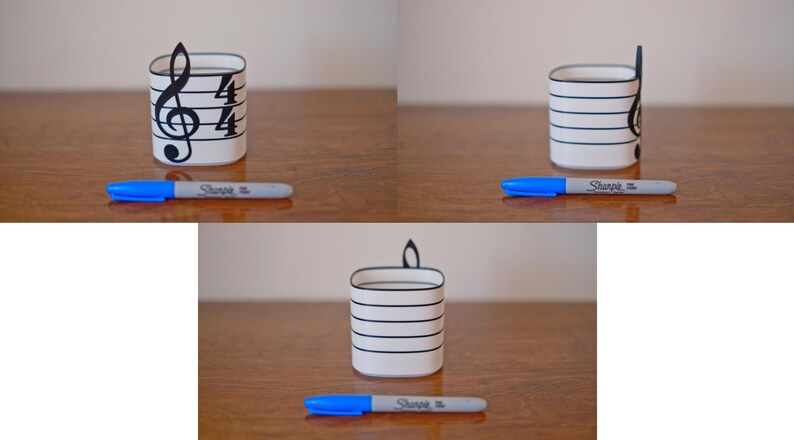 May include: A white pen holder with black horizontal lines and a treble clef design. A blue Sharpie pen rests in front of the holder. The pen holder is on a wooden surface.