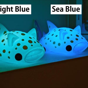 May include: Two blue, spotted, shark-shaped night lights. The light on the left is a light blue color and the light on the right is a sea blue color. The text 'Light Blue' is above the left light and the text 'Sea Blue' is above the right light.