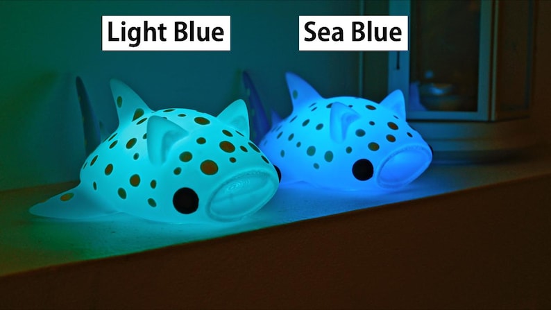 May include: Two blue, spotted, shark-shaped night lights. The light on the left is a light blue color and the light on the right is a sea blue color. The text 'Light Blue' is above the left light and the text 'Sea Blue' is above the right light.