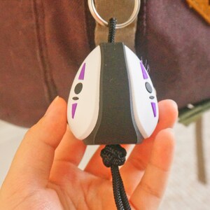 May include: A No-Face keychain, featuring a white face with black and purple details. The keychain is attached to a black cord and is held in a person's hand. The background is blurred, showing a backpack.