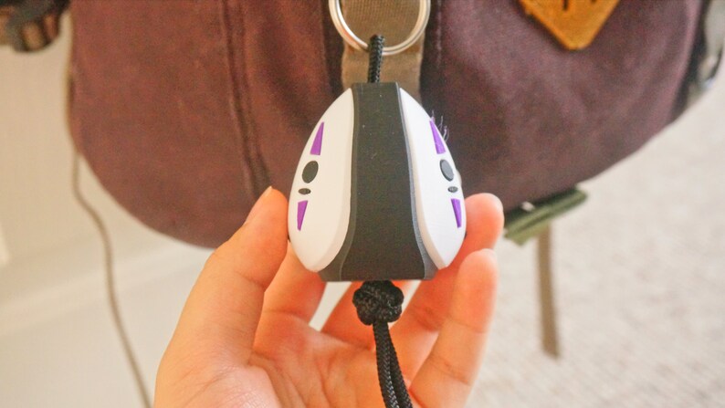 May include: A No-Face keychain, featuring a white face with black and purple details. The keychain is attached to a black cord and is held in a person's hand. The background is blurred, showing a backpack.