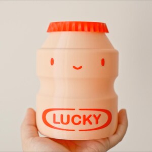 May include: A peach-colored, bottle-shaped container with a red cap. The container has a smiling face design with red eyes and a mouth. The word "LUCKY" is printed in red on the front.