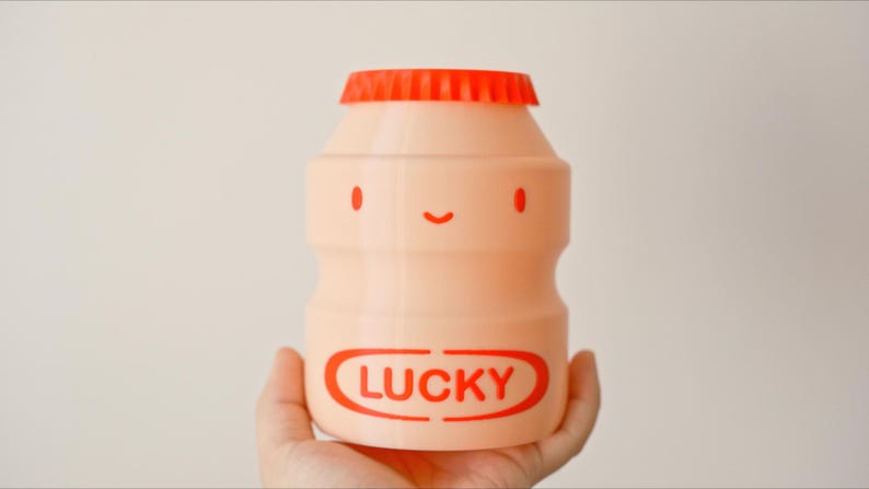 May include: A peach-colored, bottle-shaped container with a red cap. The container has a smiling face design with red eyes and a mouth. The word "LUCKY" is printed in red on the front.