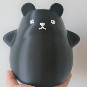 May include: A black, 3D printed, cartoon bear figurine with white eyes, nose, and mouth. The bear is standing on a hand.