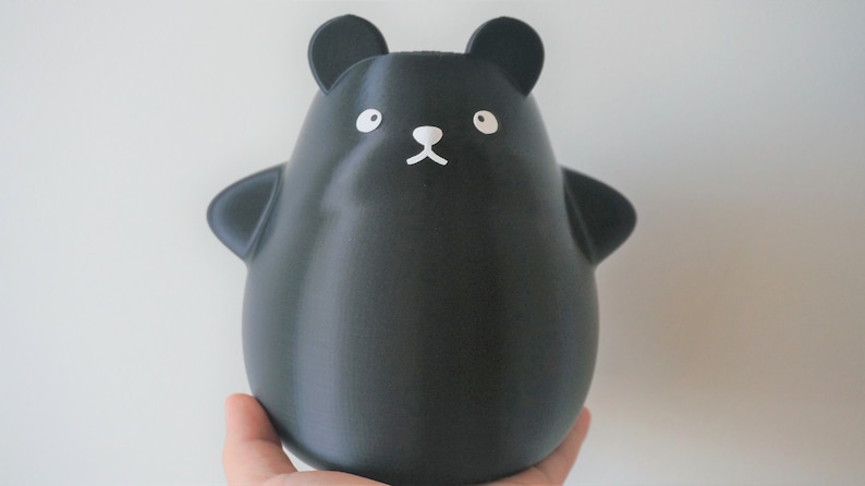 May include: A black, 3D printed, cartoon bear figurine with white eyes, nose, and mouth. The bear is standing on a hand.