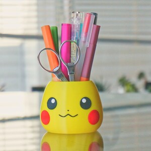 May include: A yellow Pikachu-shaped pencil holder with a red nose and black eyes. The holder is filled with colorful pens and a pair of silver scissors.