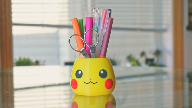 May include: A yellow Pikachu-shaped pencil holder with a red nose and black eyes. The holder is filled with colorful pens and a pair of silver scissors.