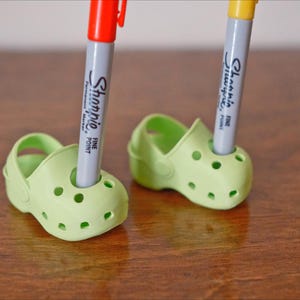 May include: Two green plastic shoes with holes, each holding a marker. The marker on the left is red and says "Sharpie Fine Point". The marker on the right is yellow and says "Sharpie Fine Point".