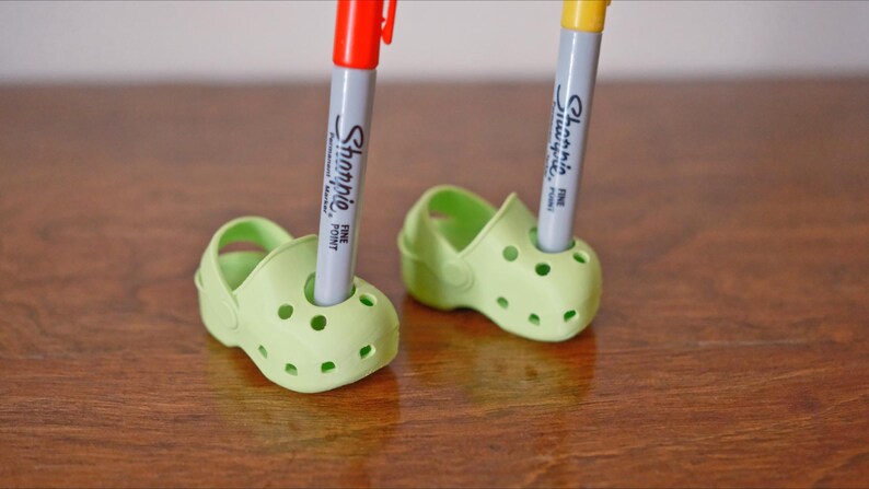 May include: Two green plastic shoes with holes, each holding a marker. The marker on the left is red and says "Sharpie Fine Point". The marker on the right is yellow and says "Sharpie Fine Point".
