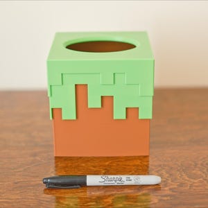 A cube tissue box cover inspired by the dirt grass block from the popular video game, Minecraft, is on a wooden table with a white background and a sharpie for size. It has bright green pixelated grass at the top half and the bottom half is brown.