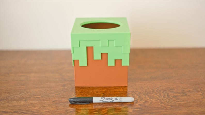 A cube tissue box cover inspired by the dirt grass block from the popular video game, Minecraft, is on a wooden table with a white background and a sharpie for size. It has bright green pixelated grass at the top half and the bottom half is brown.
