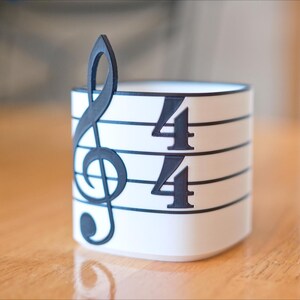 May include: A white cylindrical container with a black treble clef and 4/4 time signature design. The musical-themed decor is set on a wooden surface. The container is approximately 3 inches tall.