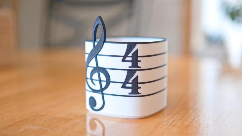 May include: A white cylindrical container with a black treble clef and 4/4 time signature design. The musical-themed decor is set on a wooden surface. The container is approximately 3 inches tall.