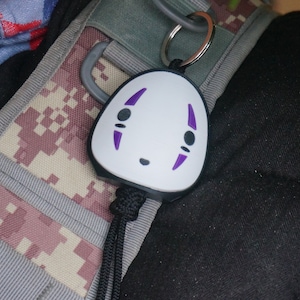 May include: A white keychain featuring a No-Face design with purple accents. The keychain has a black cord and a silver keyring. The design is a stylized face with black eyes and a small mouth.