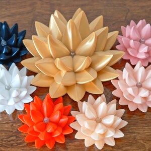 May include: Seven decorative paper flowers in various colors, including gold, white, black, pink, and orange. The flowers are arranged in a circle on a wooden surface.