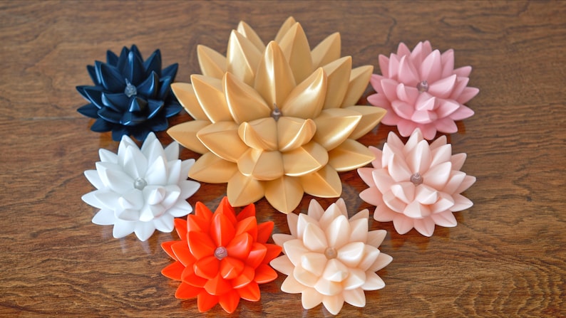 May include: Seven decorative paper flowers in various colors, including gold, white, black, pink, and orange. The flowers are arranged in a circle on a wooden surface.