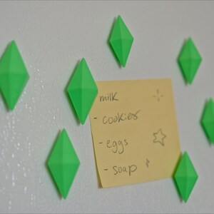 May include: A yellow sticky note with a handwritten grocery list on a white refrigerator. The list includes milk, cookies, eggs, and soap. The note is held in place by seven green diamond-shaped magnets.