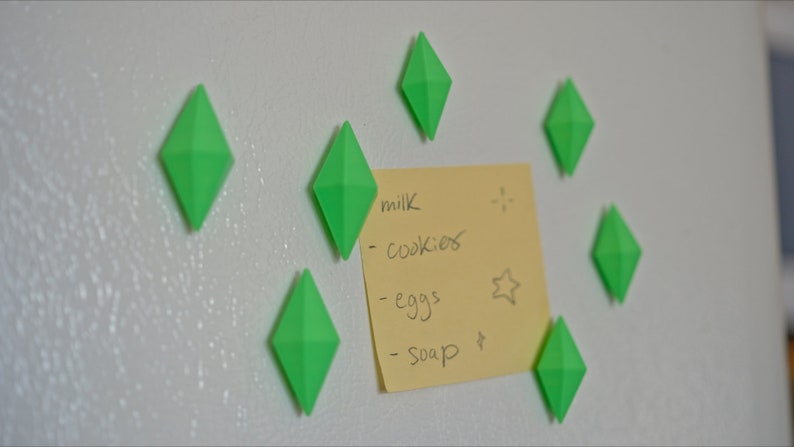 May include: A yellow sticky note with a handwritten grocery list on a white refrigerator. The list includes milk, cookies, eggs, and soap. The note is held in place by seven green diamond-shaped magnets.