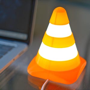 May include: An orange and white traffic cone-shaped lamp with a bright glow. The lamp is next to a laptop computer. The lamp has a square base and a pointed top. The lamp has three white stripes around the cone.