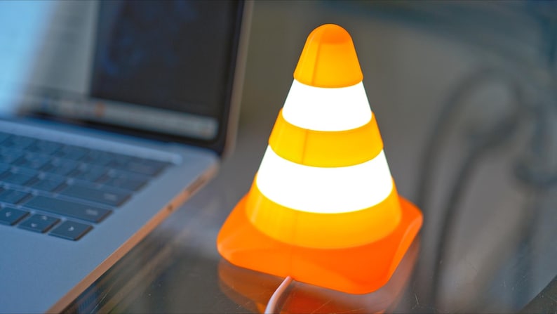 May include: An orange and white traffic cone-shaped lamp with a bright glow. The lamp is next to a laptop computer. The lamp has a square base and a pointed top. The lamp has three white stripes around the cone.