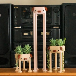 May include: Three ceramic planter pots shaped like dogs with long legs. Each pot has a different succulent plant growing inside. The pots are pink, brown, and beige. The pots are in front of a black stereo system.