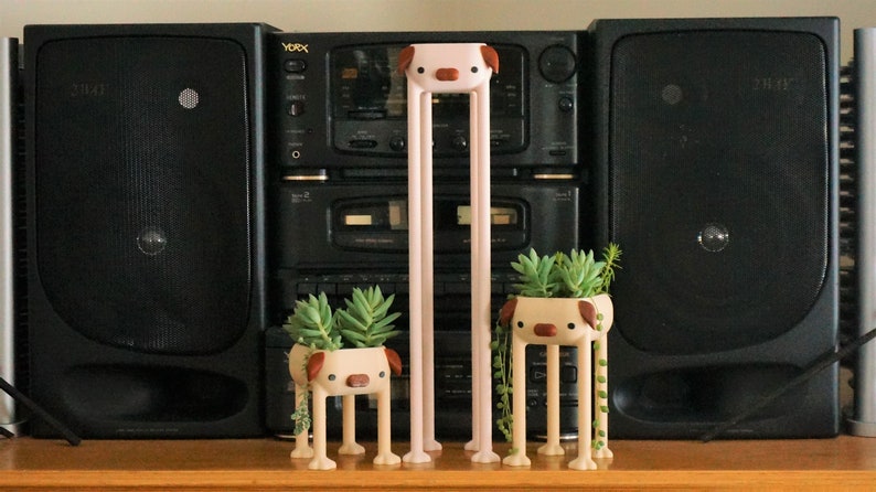 May include: Three ceramic planter pots shaped like dogs with long legs. Each pot has a different succulent plant growing inside. The pots are pink, brown, and beige. The pots are in front of a black stereo system.