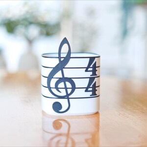 May include: A white cylindrical container with a black musical note and time signature design. The design includes a treble clef and the numbers "4/4" on the side. The container is sitting on a wooden surface.