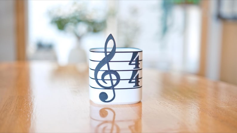 May include: A white cylindrical container with a black musical note and time signature design. The design includes a treble clef and the numbers "4/4" on the side. The container is sitting on a wooden surface.