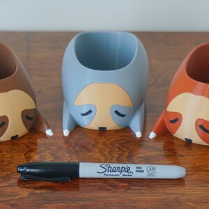May include: Three 3D printed sloth planters, one brown, one gray, and one orange, with white painted details. The planters are shaped like sloths with their heads resting on the surface. A black Sharpie fine point permanent marker is in the foreground.