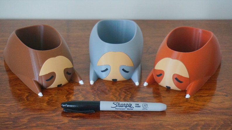 May include: Three 3D printed sloth planters, one brown, one gray, and one orange, with white painted details. The planters are shaped like sloths with their heads resting on the surface. A black Sharpie fine point permanent marker is in the foreground.