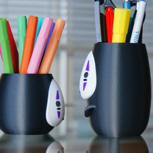 May include: Two black pen holders with a No-Face design, one filled with colorful markers and the other with pens, scissors, and a yellow highlighter. The pen holders have a white face with purple accents.