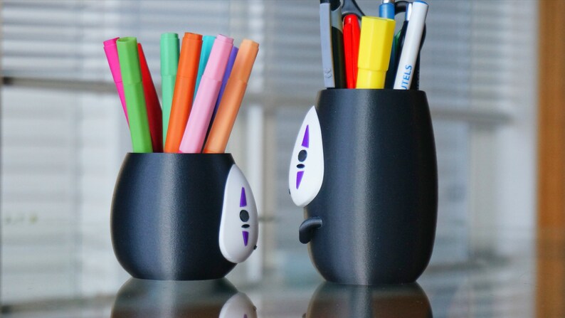 May include: Two black pen holders with a No-Face design, one filled with colorful markers and the other with pens, scissors, and a yellow highlighter. The pen holders have a white face with purple accents.