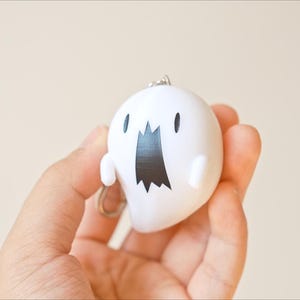 May include: A white ghost-shaped keychain with a black mouth and eyes. The keychain is held in a hand against a neutral background. The ghost has small arms and a silver keyring.