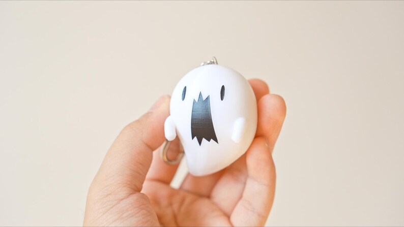 May include: A white ghost-shaped keychain with a black mouth and eyes. The keychain is held in a hand against a neutral background. The ghost has small arms and a silver keyring.