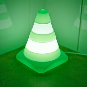 May include: A green and white LED night light shaped like a traffic cone. The cone has horizontal white stripes that illuminate. The light is on a green surface with a green background.