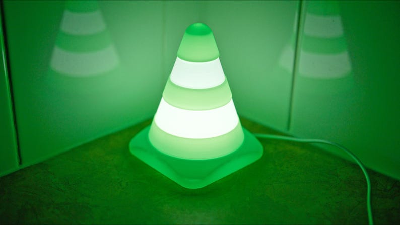May include: A green and white LED night light shaped like a traffic cone. The cone has horizontal white stripes that illuminate. The light is on a green surface with a green background.