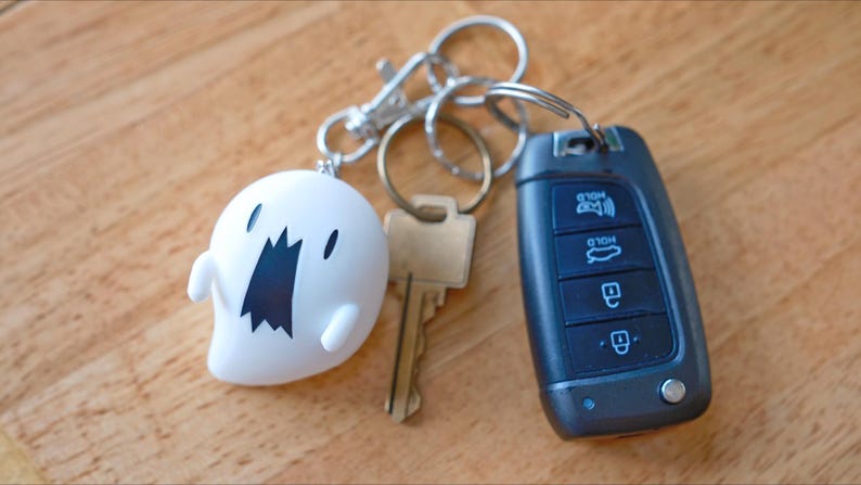 May include: A collection of keys and keychains on a wooden surface. Includes a white ghost-shaped keychain with a black mouth, a car key fob, and a standard metal key.