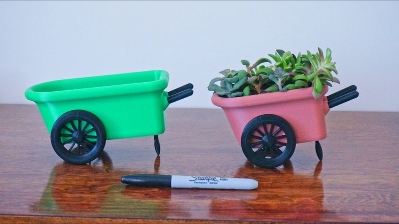 May include: Two miniature wheelbarrows, one green and one pink, with black wheels and handles. The pink wheelbarrow has a small succulent plant inside.