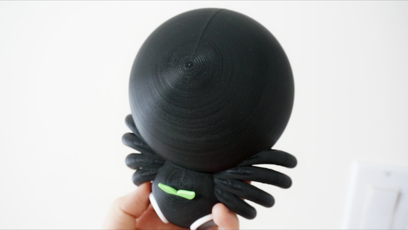May include: Black and green 3D printed spider figurine with a round head and eight legs.