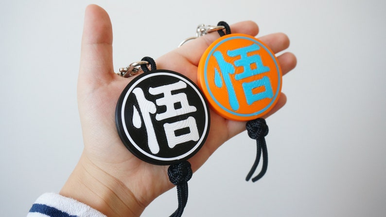Puede incluir: Dos llaveros redondos con el car&aacute;cter chino "信" (que significa "confianza") en blanco sobre fondo negro y azul sobre fondo naranja.
