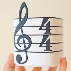 May include: A white cylindrical container with a black treble clef and 4/4 time signature design. The musical symbol is on the left side, and the time signature is on the right. The container is held by a person.